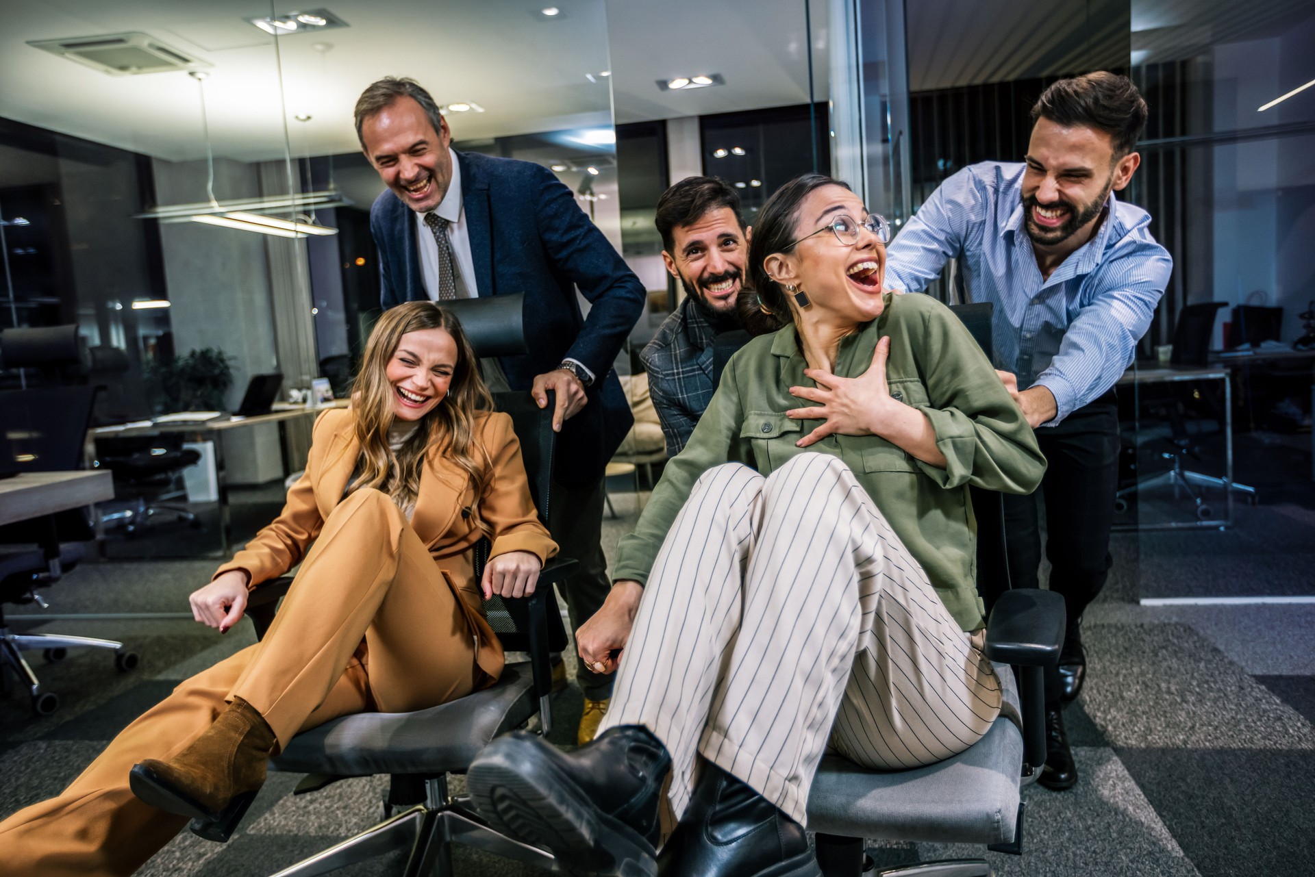 Playful scene in a modern office with businesspeople engaging in a friendly competition. In the image, colleagues are playfully pushing each other on office chairs